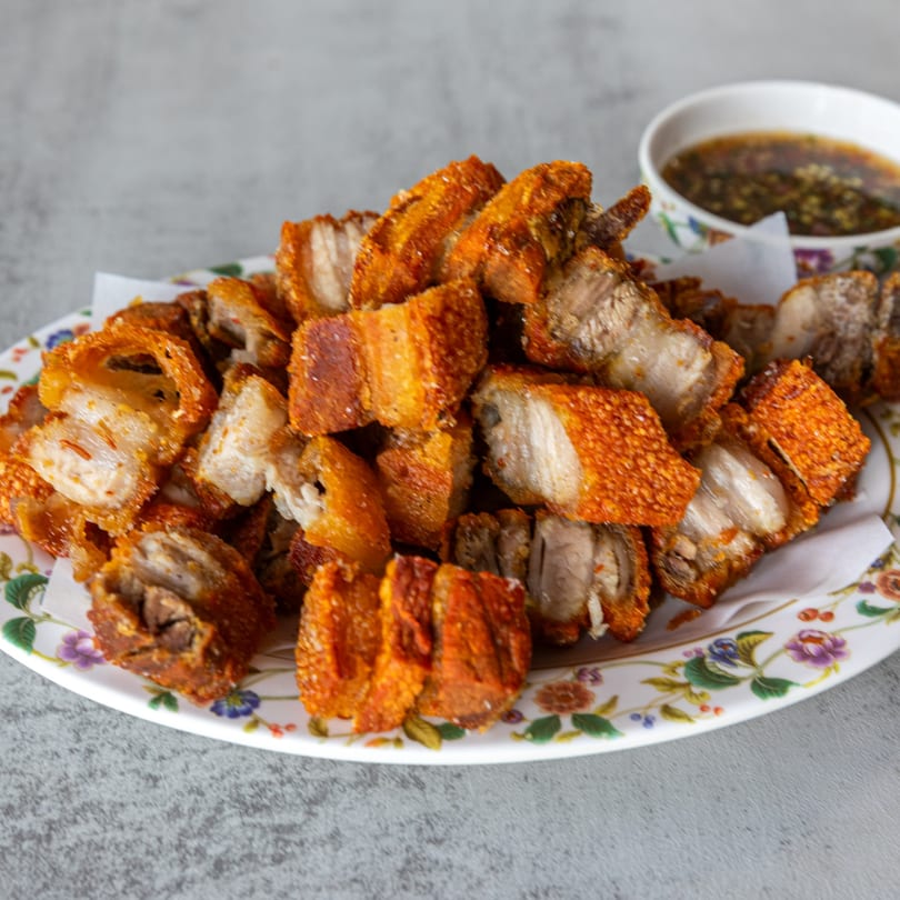 Crispy fried pork belly pieces with golden skin on a floral plate, with a dipping sauce in the background.