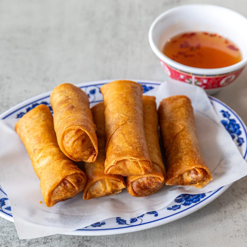 Golden-brown crispy spring rolls on a white plate with blue floral trim, served with dipping sauce.